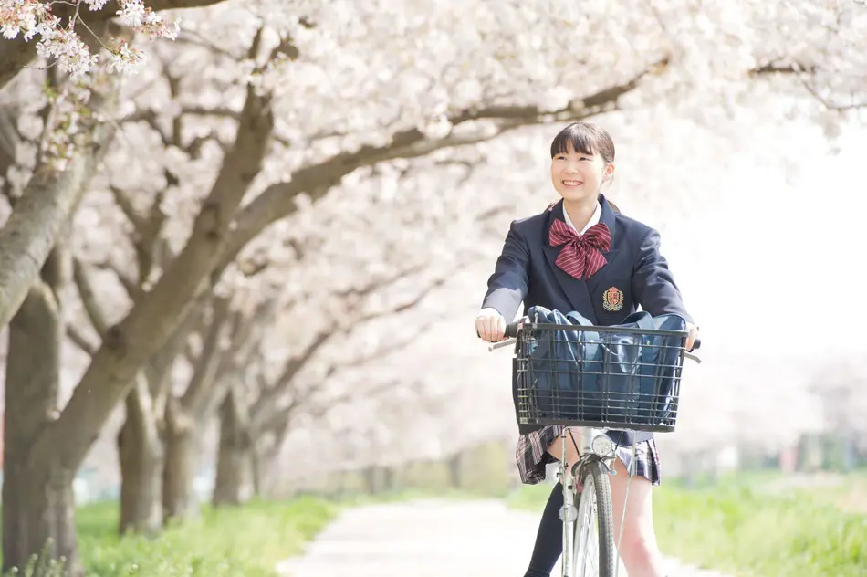 春の桜並木の下を自転車で走る中高一貫校の女子生徒